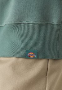 Närbild av grön ribbad sweatshirt-kant med Dickies-logotyp på en tag ovanför beige byxor.