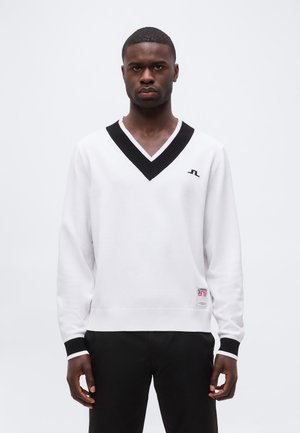 Mand iført hvid V-hals sweater med sort kant og sorte bukser, stående mod en ensfarvet hvid baggrund.