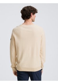 Beige, hosszú ujjú pulóver kerek nyakkivágással és bordás mandzsettával. Puha tapintású, sima felületű, és egy kis logó részlet található az ujján.