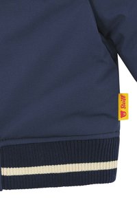 Giacca blu navy con finitura strutturata, polsini a coste con una striscia color crema e un'etichetta gialla con un logo di un orso.