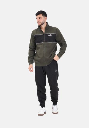 Hombre vistiendo una chaqueta con cremallera de Puma en verde oliva y negro, pantalones joggers negros de Puma y zapatillas blancas, posando contra un fondo liso.