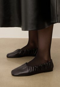 Bailarinas de piel negra con una textura fruncida alrededor de la abertura, suela plana, combinadas con medias opacas negras y una falda de tela negra.