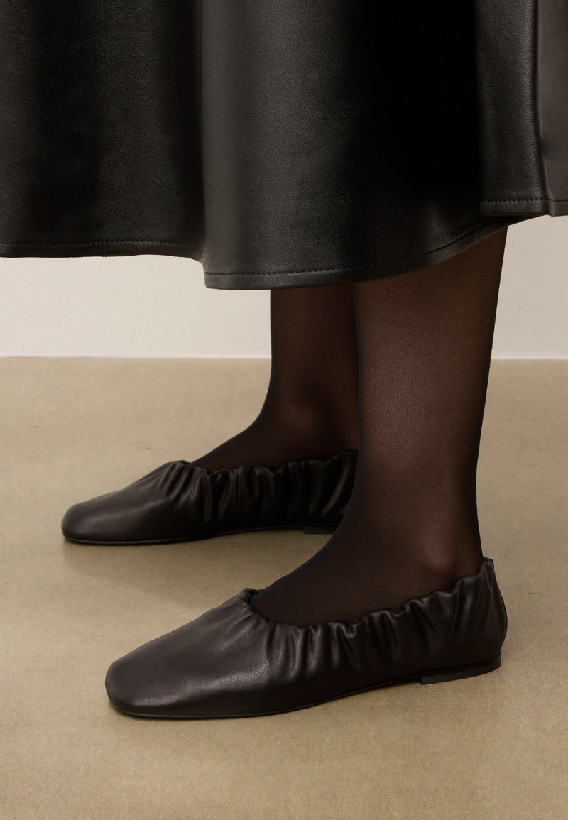 Bailarinas de piel negra con una textura fruncida alrededor de la abertura, suela plana, combinadas con medias opacas negras y una falda de tela negra.