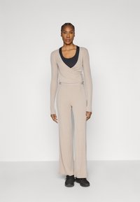 Beżowa bluzka kopertowa z długimi rękawami i wiązaniem, zestawiona z szerokimi spodniami. Pod spodem czarna koszulka na ramiączkach. Modelka stoi z rękami wzdłuż ciała, ma na sobie czarne buty.