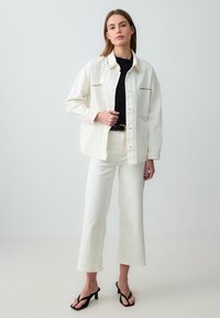 Chemise en denim blanche avec des boutons à pression et des coutures noires contrastées, associée à un jean flare blanc et des sandales à talons noires.