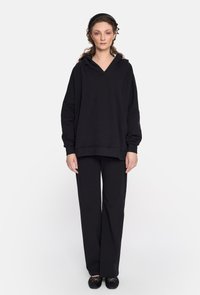 Czarna bluza z kapturem o luźnym kroju, zestawiona z dopasowanymi luźnymi spodniami, obie wykonane z miękkiego materiału. Nosi się z czarnymi płaskimi butami.