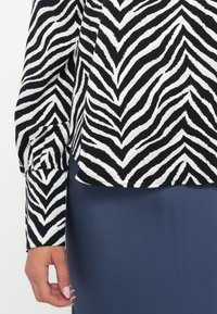 Main droite d'une femme avec des ongles rose pâle à côté d'un haut à manches longues noir et blanc à imprimé zèbre et d'une jupe bleu marine.