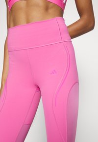 Zbliżenie na osobę w jasnoróżowych, wysokich legginsach sportowych z delikatnym logo Adidas oraz pasującym stanikiem sportowym.