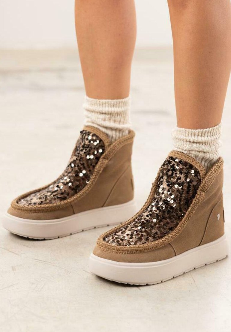 Botas de tobillo de ante marrón con acentos de lentejuelas, con una suela blanca y un ribete trenzado alrededor de la abertura. Calcetas de lana texturizadas y cálidas a la vista.
