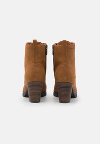 Bottines en daim marron avec un talon en bois empilé, présentant une fermeture éclair latérale et une boucle à l'arrière pour un enfilage facile. Texture lisse.