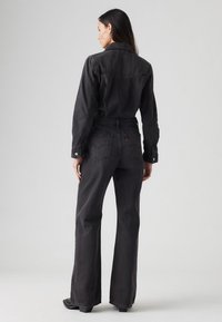 Juodas džinsinis šokinys su ilgomis rankovėmis, prigludusia juosmens linija, plačiais kelnais ir mygtukų detalėmis, turintis subtilų išblukusį tekstūrą ir galines kišenes.