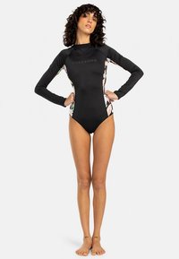 Schwarzer Rashguard mit langen Ärmeln und floralen Seitenpartien, aus dehnbarem Material gefertigt. Auf der Vorderseite befindet sich ein dezenter Logoaufdruck.