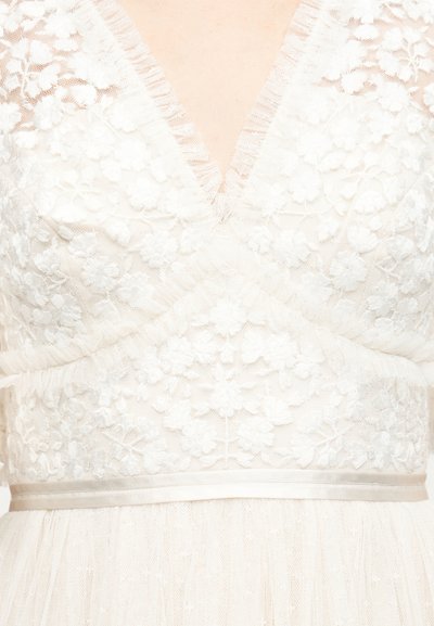 Λευκό δαντελένιο ένδυμα με λουλουδένια σχέδια, ντεκολτέ σε σχήμα V και διάφανο ύφασμα γύρω από τη μέση και τους ώμους.