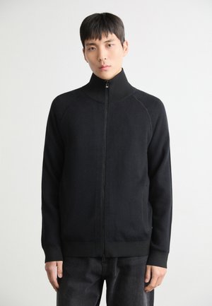 Felpa nera con zip e colletto alto, realizzata in tessuto morbido, con maniche raglan e polsini a coste, indossata con jeans scuri.