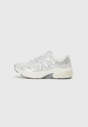 Grey athletic shoe with metallic accents, mesh upper, plush sole, textured detailing, and rounded toe design. Features laces and branding.