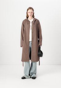 Long brown coat with a hood, button closure, and side ties, paired with a light sweater and wide-leg jeans. Black bag in hand.