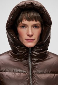 Pruun paks jakk suure kapuutsi ja kõrge kraega; esil on tõmblukk ning sile, läikiv tekstuur.