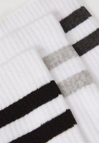 Trzy pary skarpet w paski: białe z czarnymi, szarymi i czarnymi paskami. Miękki materiał z prążkowaną teksturą. Minimalistyczny design.