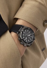 Czarny zegarek z teksturowanym czarnym paskiem z gumy, sześciokątną srebrną kopertą i czarną tarczą z srebrnymi akcentami oraz funkcjami chronografu.