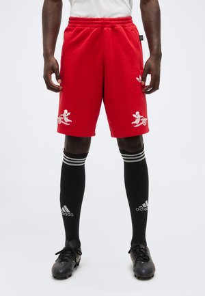 Bărbat purtând pantaloni scurți roșii Adidas cu designuri albe de heruvim și tun, șosete negre Adidas până la genunchi și ghete negre, stând pe un fundal alb.