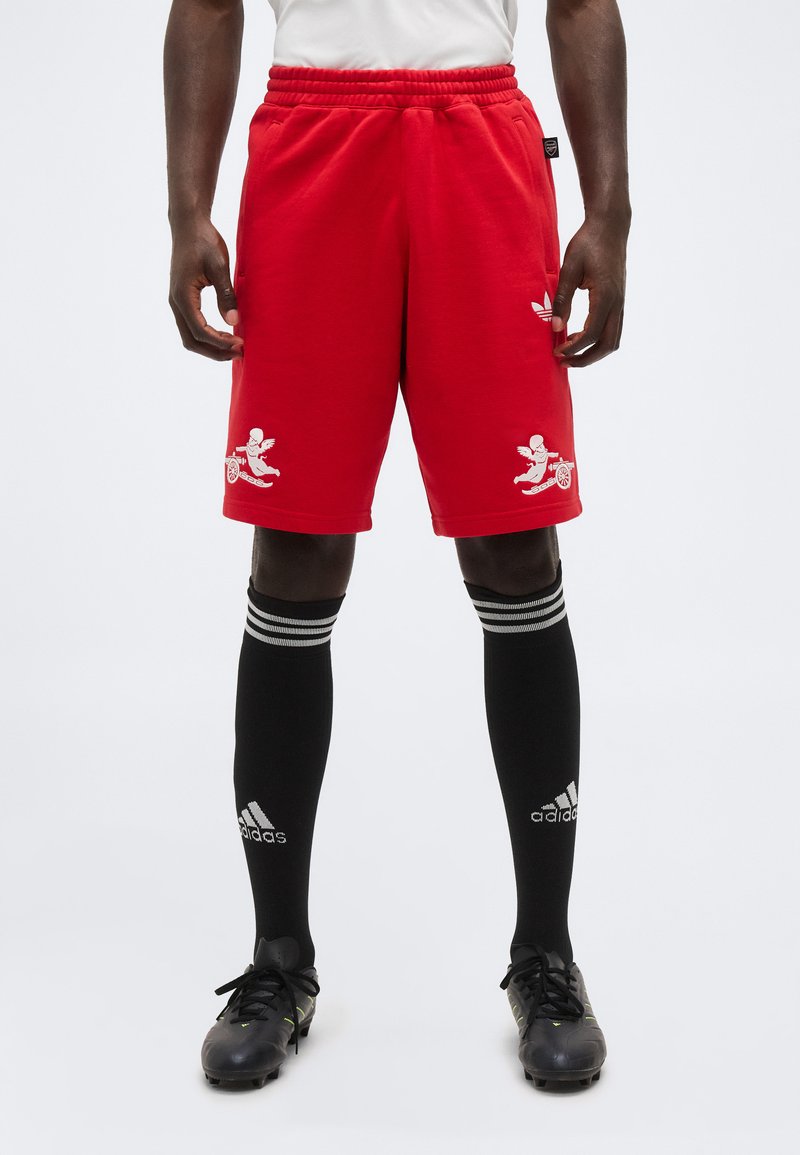 Muž oblečený v červených kraťasoch Adidas s bielymi motívmi cherubína a dela, čierne vysoké Adidas ponožky a čierne kopačky, stojaci na bielom pozadí.