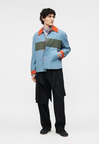 Άνδρας με μπουφάν σε μπλοκ χρωμάτων με μπλε, πράσινες και πορτοκαλί λεπτομέρειες, συνδυασμένο με μαύρο παντελόνι με στρώσεις και μαύρα παπούτσια, κοιτάζοντας προς τα πλάγια.