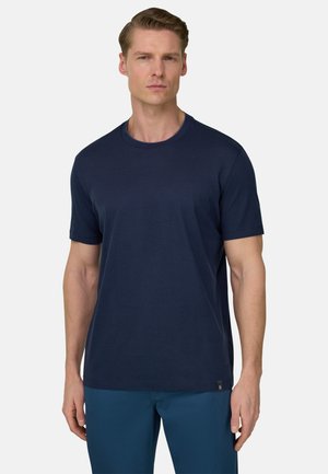 Mann steht frontal, trägt ein marineblaues T-Shirt mit kurzen Ärmeln und blaue Hosen vor einem schlichten hellen Hintergrund.