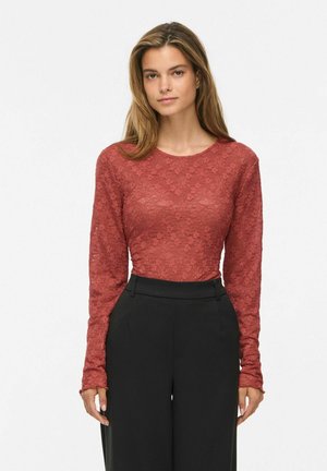 Femme aux cheveux châtain clair portant un top en dentelle rouge à manches longues et un pantalon noir taille haute, debout devant un fond blanc.