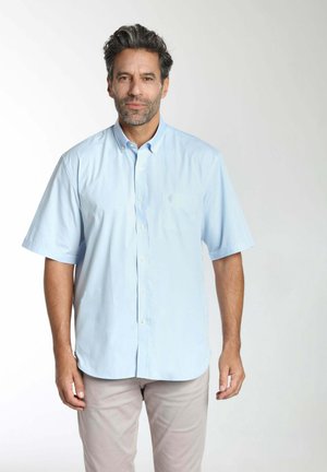 Homme aux cheveux foncés et à la barbe portant une chemise à manches courtes bleu clair à boutons et un pantalon beige, debout devant un fond uni.
