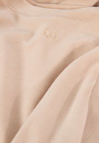 Lichtbeige stof met een gladde textuur, voorzien van een klein geborduurd logo in de linker bovenhoek. Het kledingstuk is iets gekreukt.