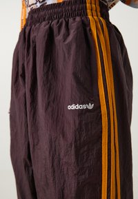 Purpurinės sportinės kelnės su elastinga juosta, oranžinėmis šoninėmis juostelėmis ir sidabriniu Adidas logo kairėje kojoje. Lengvas, tekstūruotas audinys.