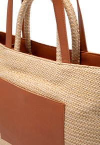 Tkaná slaměná taška s hnědými koženými akcenty. Má dvojité uši, obdélníkovou koženou kapsu vpředu a pevný tvar.