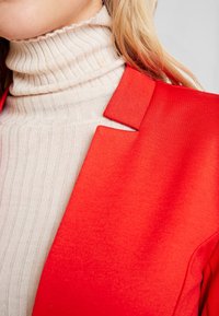 Blazer rouge avec un col structuré, associé à un pull à col roulé côtelé beige. La texture lisse du blazer contraste avec le tissu du pull.