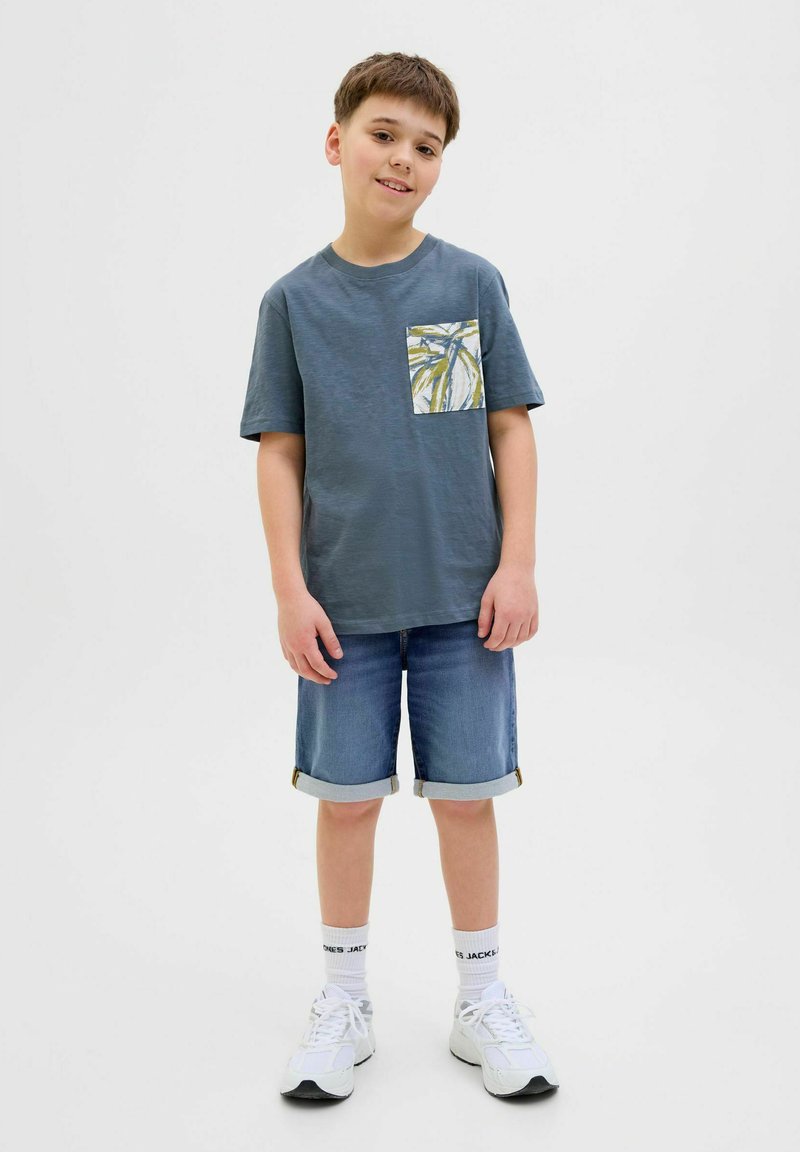 T-shirt gris à manches courtes avec une poche à motifs, associé à un short en denim bleu. Des baskets blanches et des manches retroussées complètent le look.