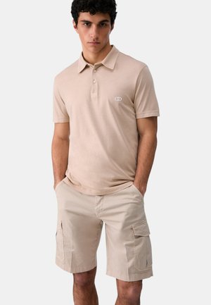 Giovane uomo con polo beige chiaro e pantaloncini cargo abbinati, in piedi con le mani nelle tasche su sfondo semplice.