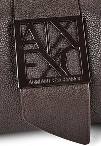Σκούρο καφέ δερμάτινη υφή με γυαλιστερή μαύρη πλακέτα λογότυπου Armani Exchange, τοποθετημένη σε λουράκι.