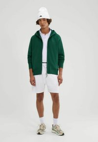 Sudadera verde con cremallera, polo de manga corta blanco, pantalones cortos blancos, gorro blanco y zapatillas multicolores. Tela suave y corte casual.