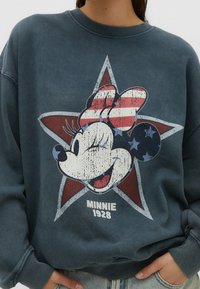 Donkerblauwe trui met een verweerde afbeelding van een glimlachend figuurtje met een rood-wit gestreept strikje, geplaatst binnen een ster met details.