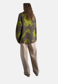 Personne aux cheveux longs et ondulés portant un pull oversize à motifs marron et verts et un pantalon beige ample, vue de dos sur un fond blanc.