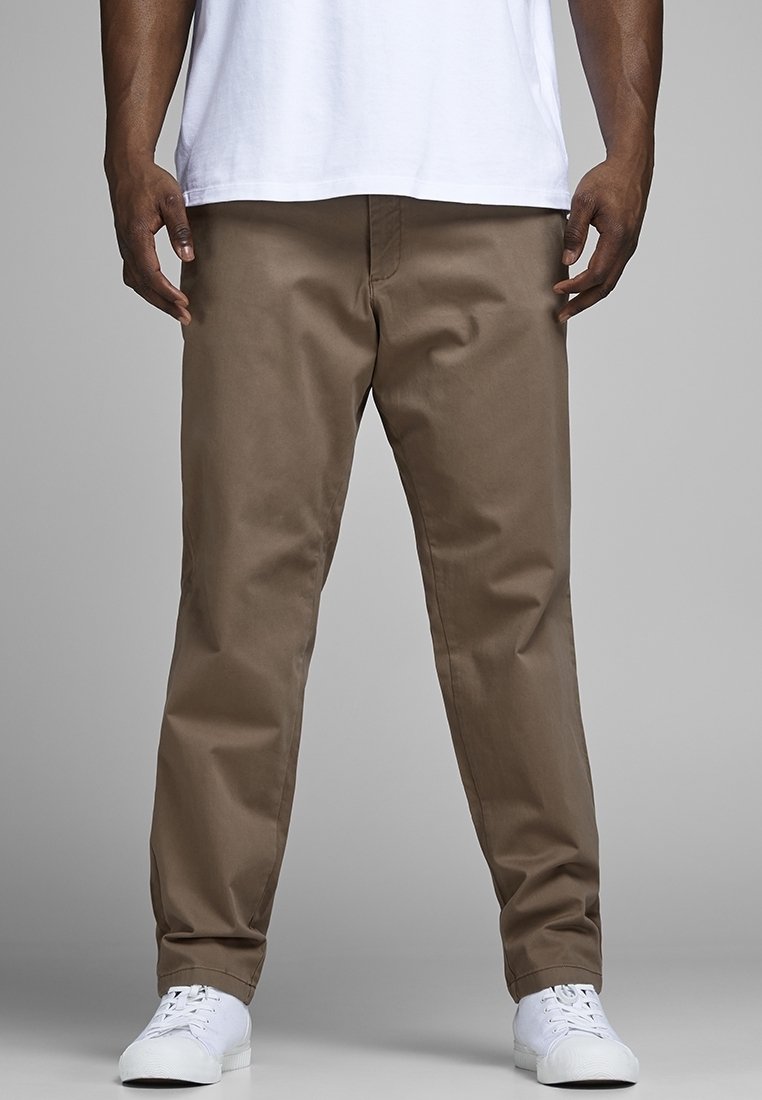 Homme portant un pantalon droit marron, des baskets blanches et une chemise blanche, debout devant un fond gris neutre.
