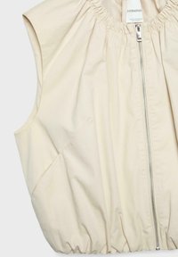 Veste beige sans manches avec encolure élastiquée froncée, fermeture éclair frontale, poches latérales, et étiquette "Stradivarius" à l'intérieur du col.