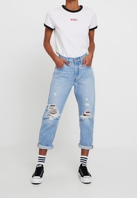 T-shirt blanche à manches courtes avec des accents noirs et un logo rouge, associée à un jean clair délavé et des baskets noires avec des semelles blanches.