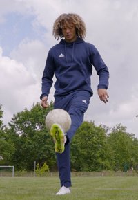 Idrettsutøver i marineblå hettegenser og bukser, sparker på en grå fotball. Grønne og rosa detaljer på fottøyet, med en gresslette og trær i bakgrunnen.