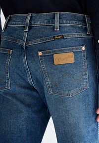 Widok z tyłu niebieskich dżinsów Wrangler z widocznym przeszyciem, miedzianymi nitami i brązową skórzaną łatką z logo na prawym tylnym kieszeni.