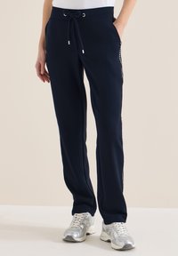 Pantalon bleu marine à cordon avec poche latérale, bande blanche à motif sur les jambes, porté avec des chaussures de sport argentées, debout devant un fond uni.