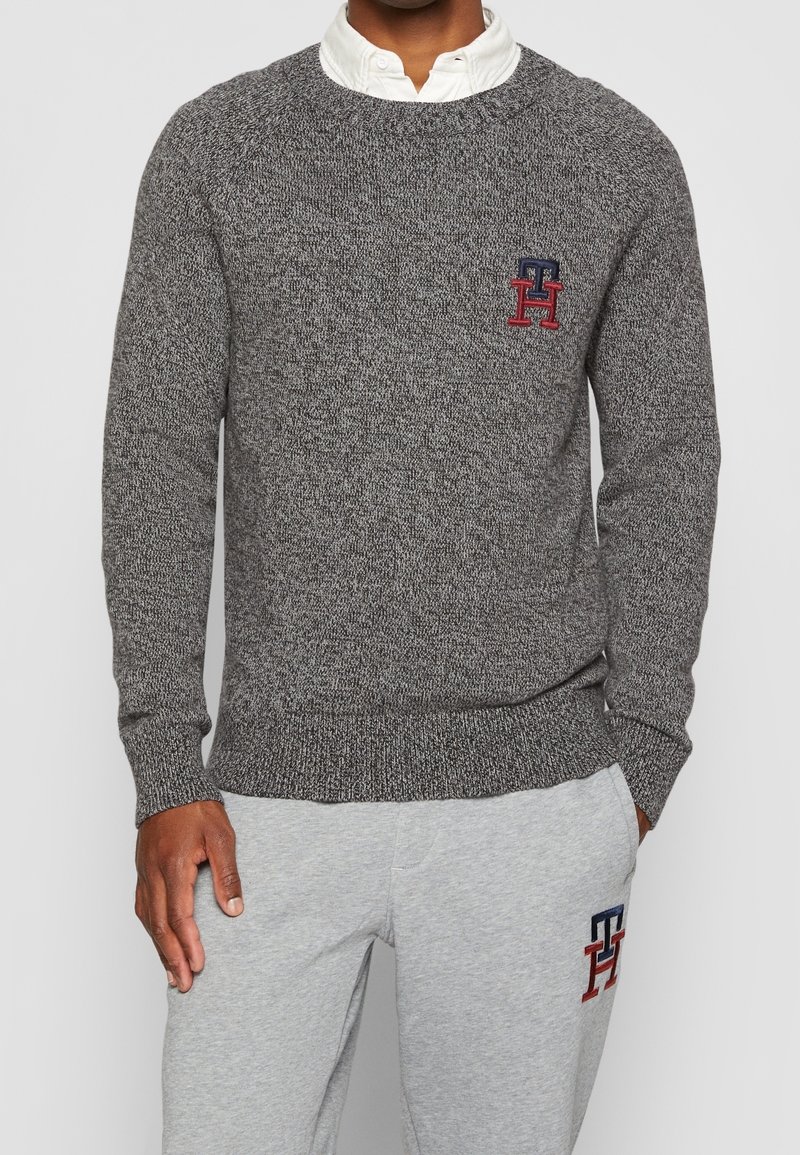 Grauer Strickpullover mit Rundhalsausschnitt, gerippten Bündchen und Saum. Mit einem kleinen roten und blauen Logo auf der linken Brust.