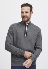 Pulover gri cu guler înalt, având un fermoar pe jumătate cu dungi de accent roșie și albastră. Mansete și tiv cu nervuri; material tricotat texturat.