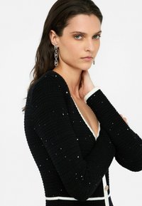 Abito nero con texture, scollatura a V, rifiniture bianche e dettagli gioiello. Le maniche lunghe e il bottone decorativo esaltano il suo design strutturato.