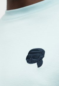 Ανοιχτό μπλε πουκάμισο με ένα μικρό μαύρο κεντητό σήμα στο στήθος που απεικονίζει ένα στυλιζαρισμένο πρόσωπο με γυαλιά ηλίου και μαντήλι στο κεφάλι.