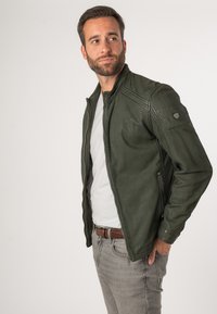 Olijfgroene leren jas met ritssluiting, schouderstiksels en zijzakken. Draag je over een lichtgrijs t-shirt en lichtgrijze jeans.